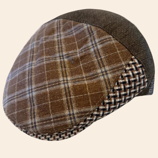 A brown plaid flat cap with a dark brown woven side panel and a textured brown fabric crown, resting on a crumpled white surface.