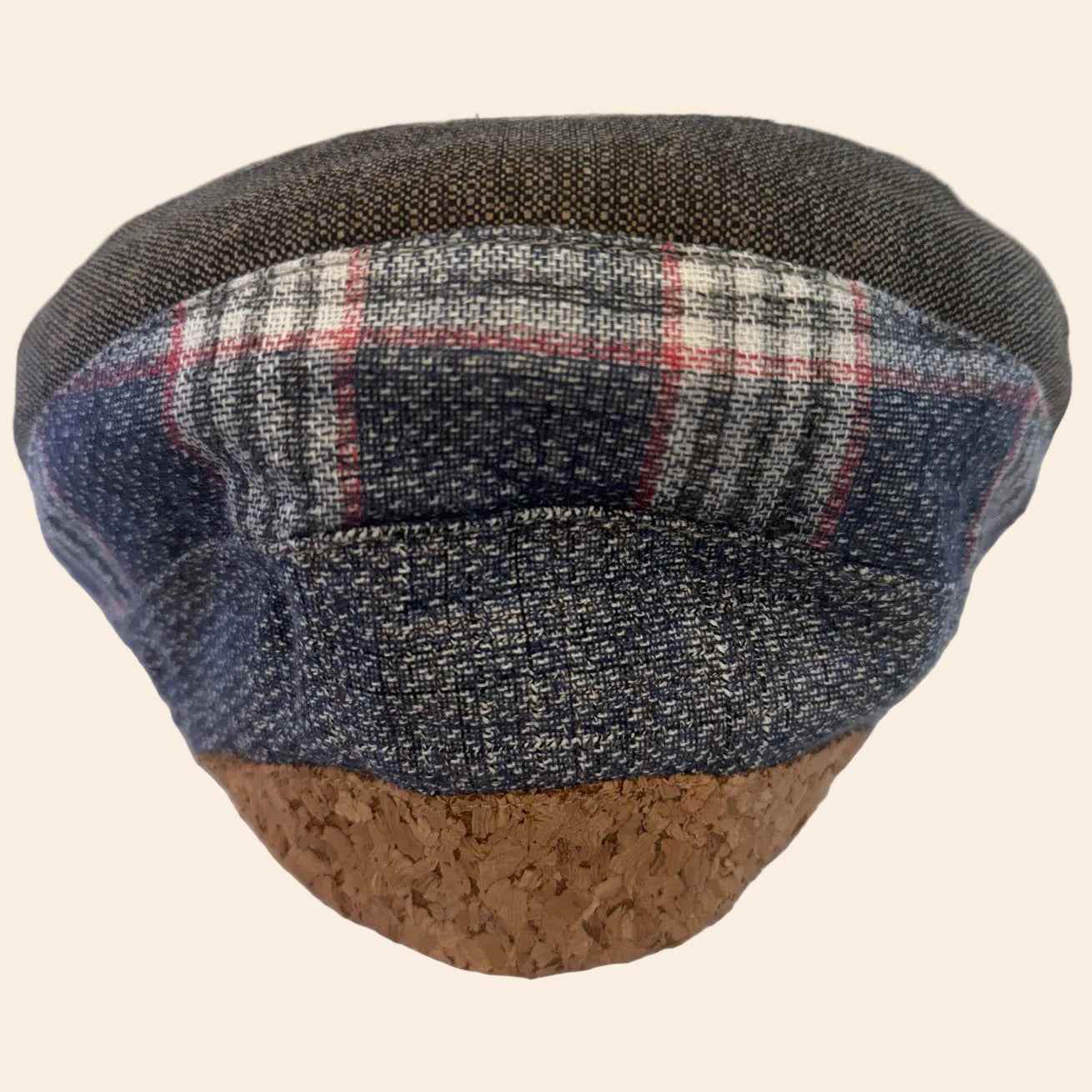A low-profile flat cap hat featuring a textured dark gray and navy woven fabric top, a plaid pattern band in gray, white, and red, and a natural cork base.
