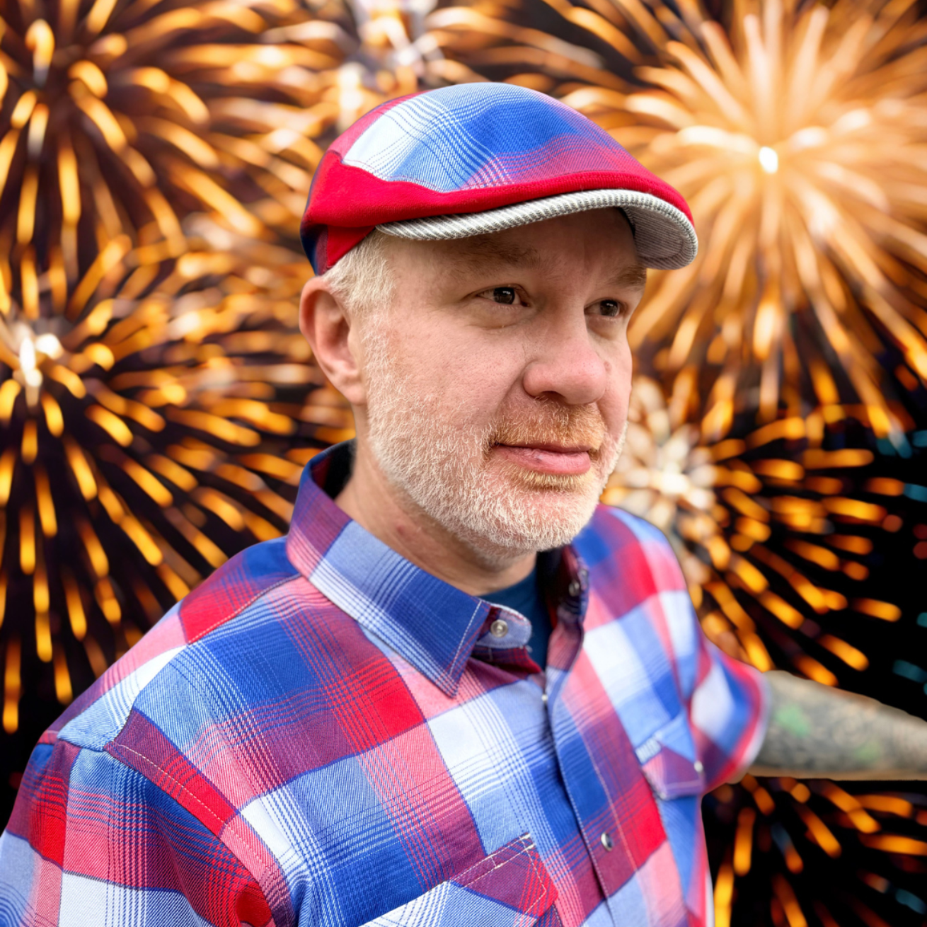 Man wearing a red, white, and blue cap and plaid shirt with fireworks in the background