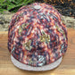 A corduroy flat cap with a multicolor abstract pattern in red, orange, blue, and beige, featuring a contrasting light gray knit brim, displayed on a wooden surface with outdoor foliage in the background.