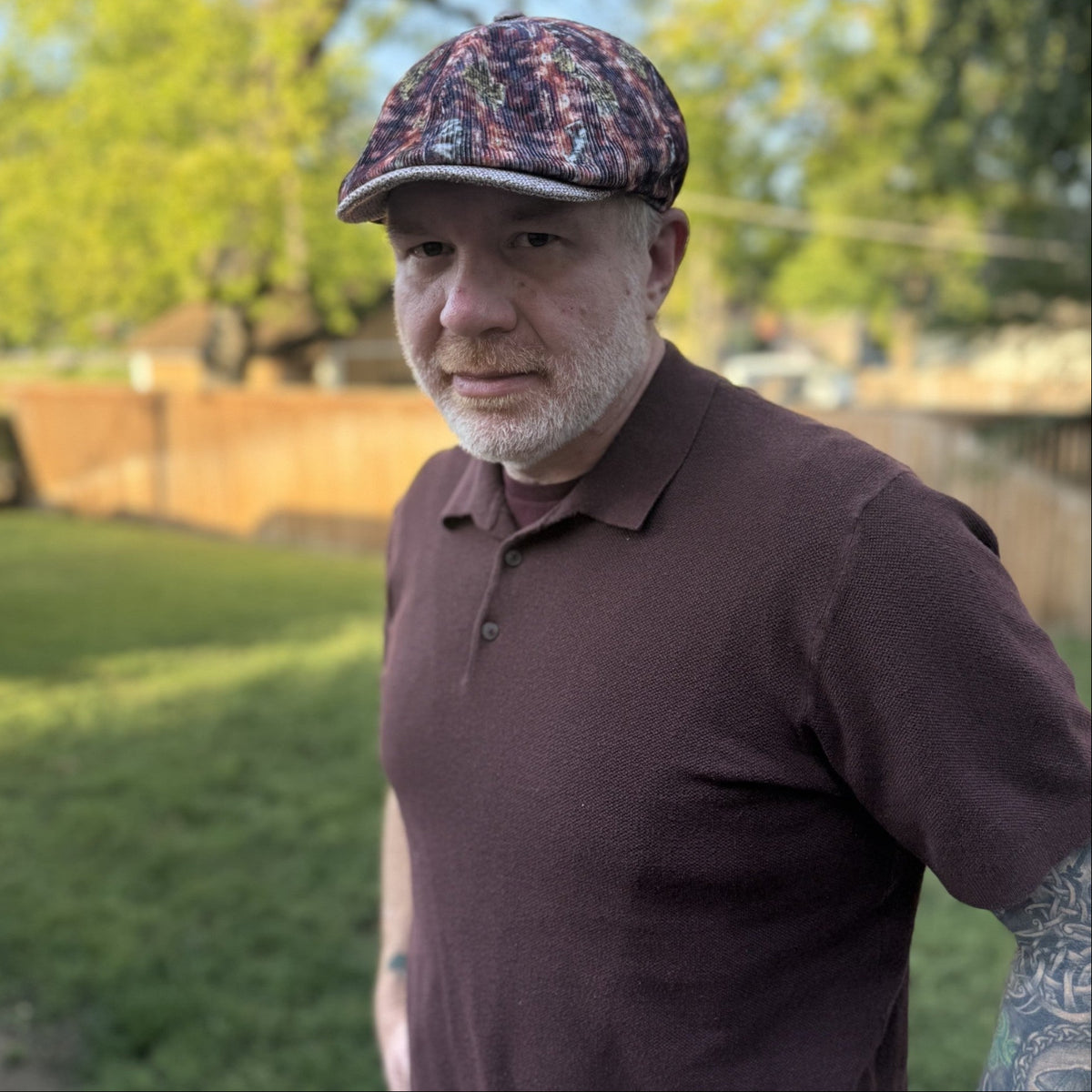 A man wearing a brown polo shirt and a patterned flat cap, standing outdoors in a grassy backyard.