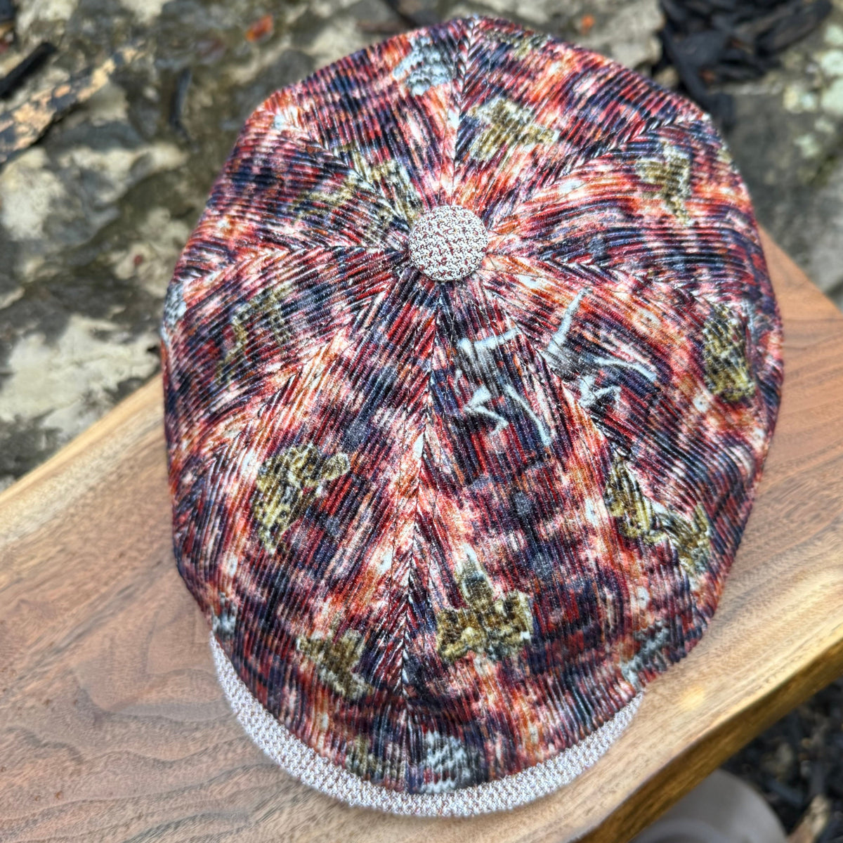 A textured, multi-colored flat cap with a floral or starburst pattern in red, orange, black, and gold, resting on a wooden surface outdoors.