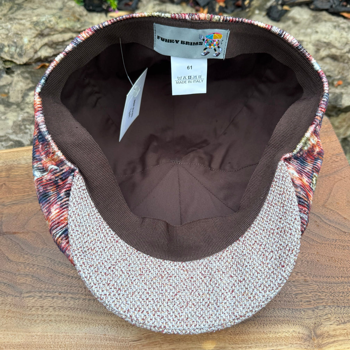 A vintage-style flat cap with a brown ribbed knit brim and a multicolor tweed patterned crown, shown on a wooden surface with a rocky background.