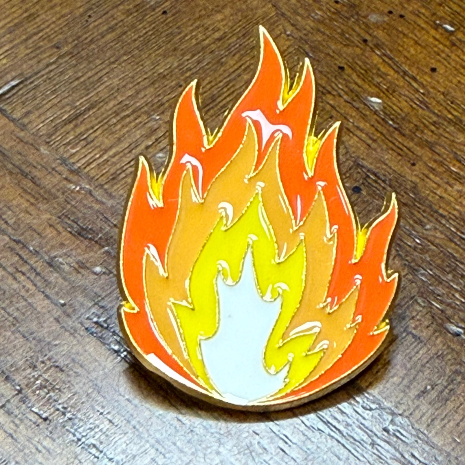 A small enamel pin shaped like a flame, shown on a dark wood surface. The pin has layered flame colors including orange, yellow, and white, outlined in gold-toned metal.