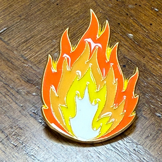 A small enamel pin shaped like a flame, shown on a dark wood surface. The pin has layered flame colors including orange, yellow, and white, outlined in gold-toned metal.
