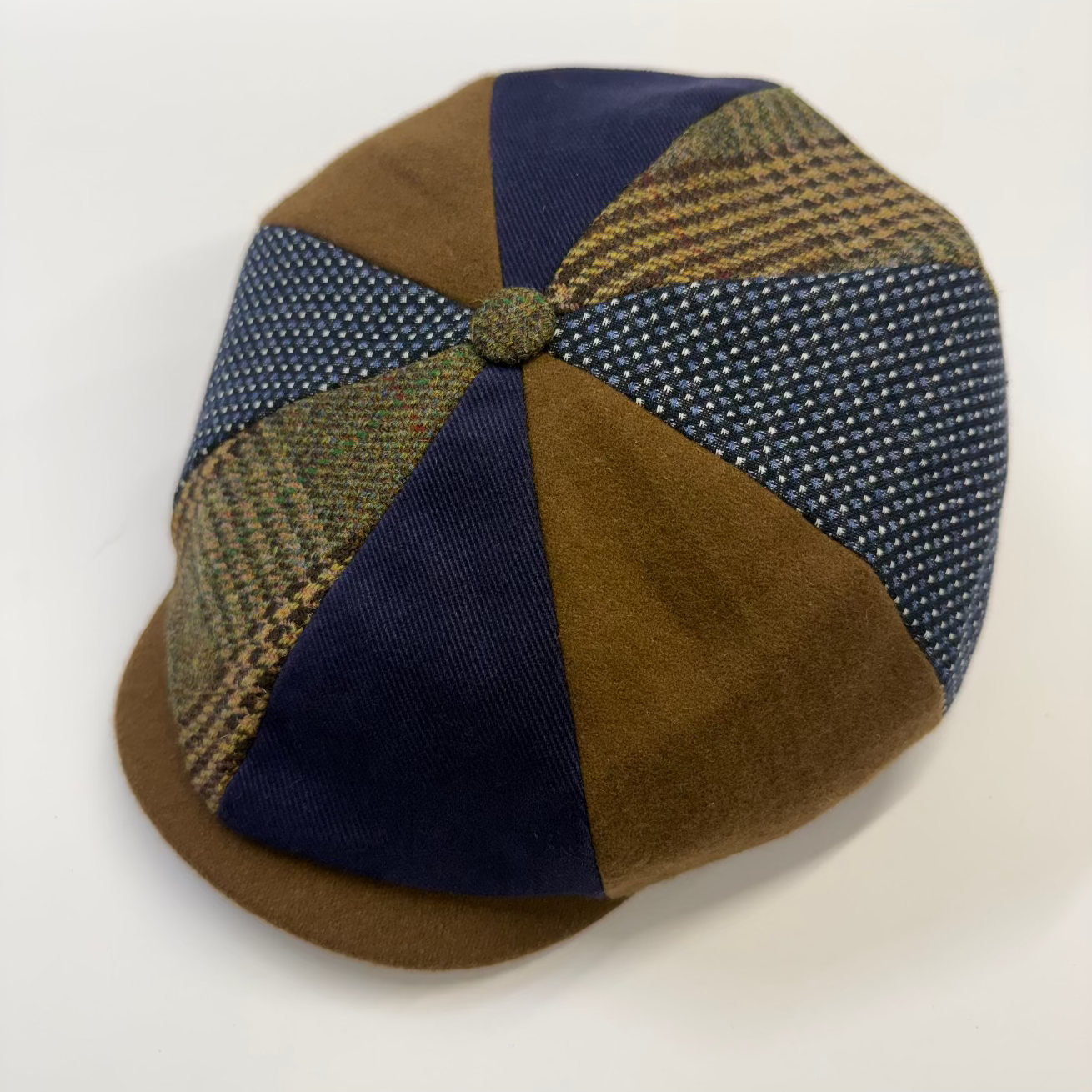 A vintage-style flat cap made of patchwork fabrics, featuring brown suede-like panels, navy blue solid panels, and tweed plaid panels in green and brown, with a matching button on top, set against a plain white background.