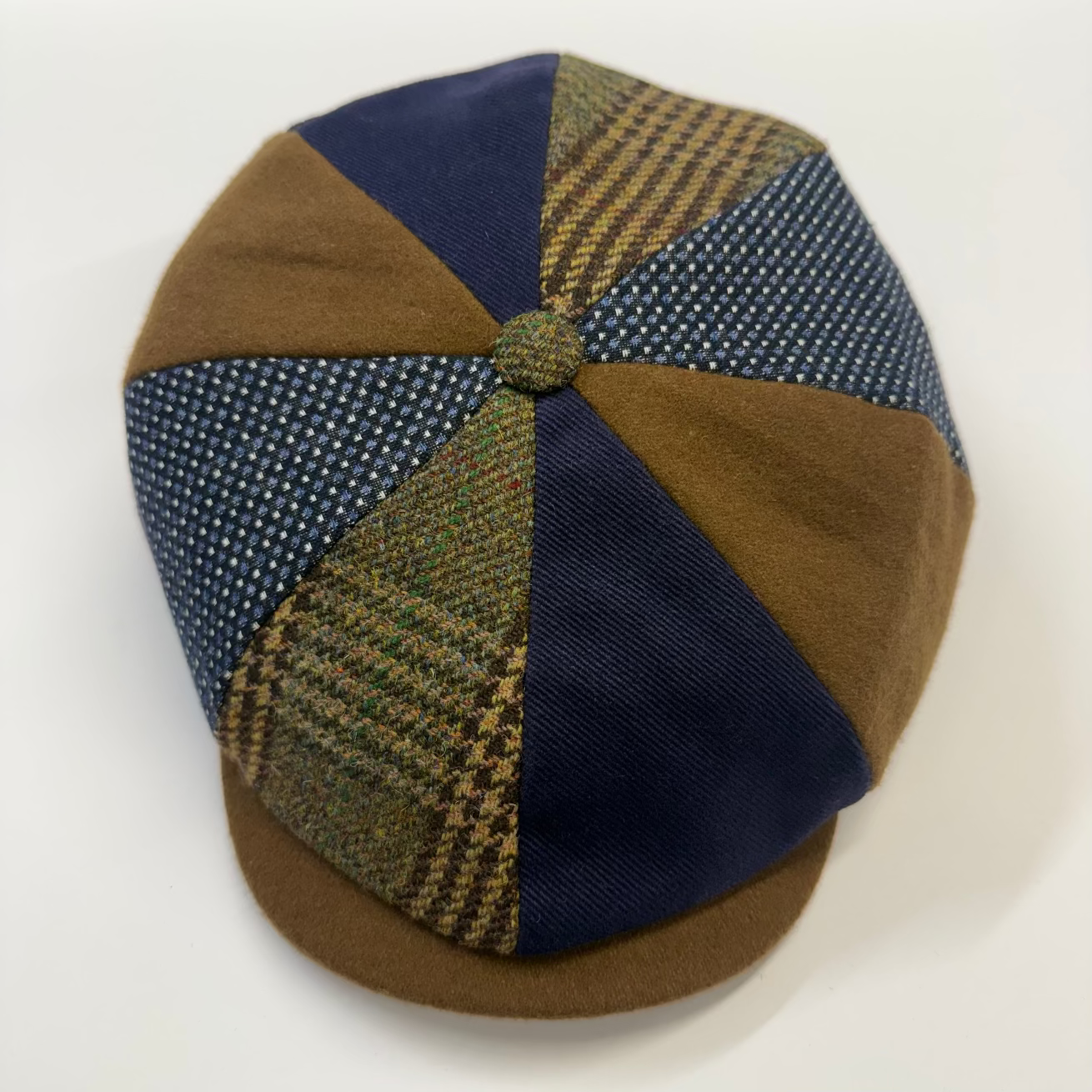 A handmade flat cap made of multiple fabric panels, featuring navy blue solid, brown wool, and green-and-brown houndstooth tweed sections, with a matching brown visor and button.