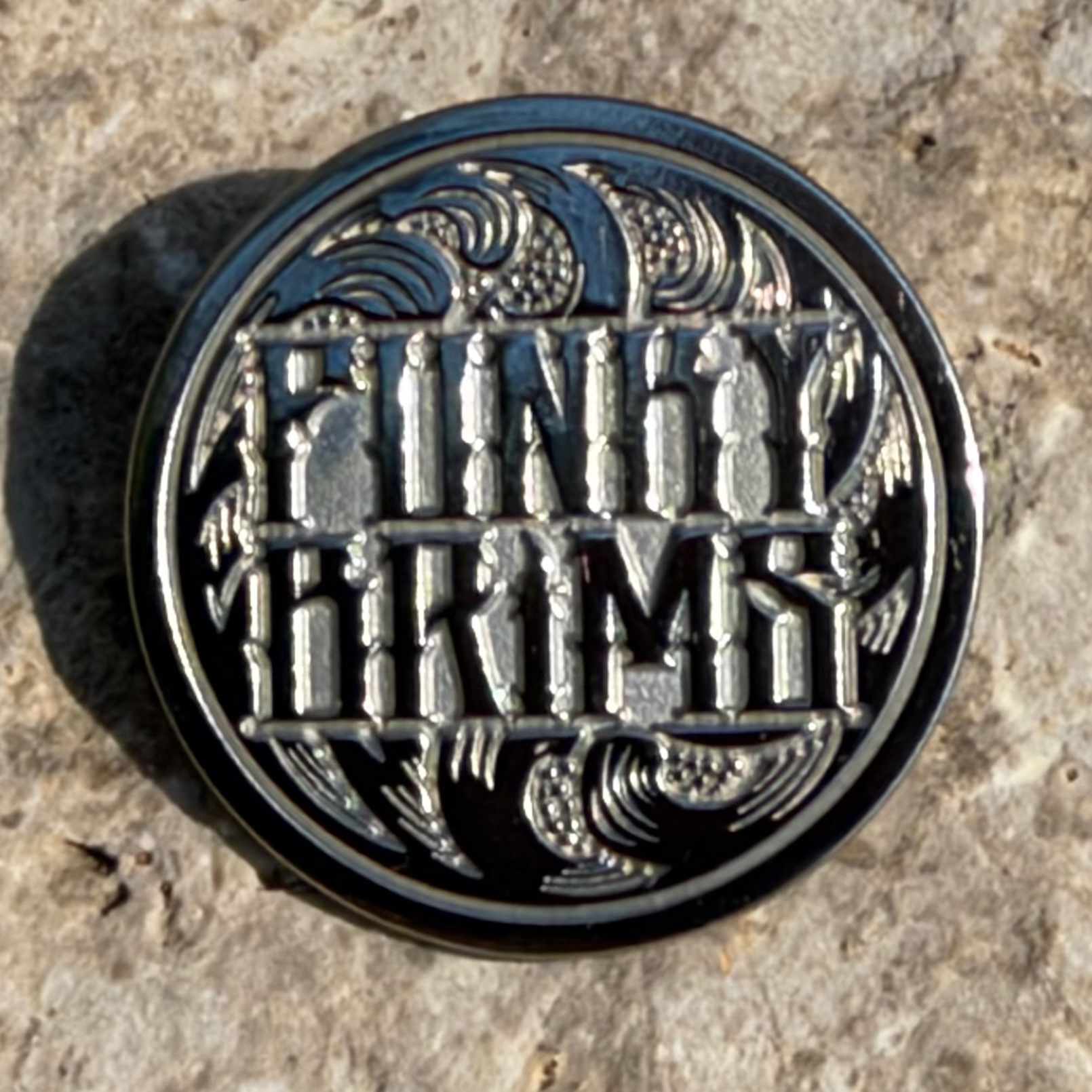 A round black and silver metal pin featuring the text 'FINGY ARMY' in stylized lettering with decorative wave and figure motifs, photographed on a textured stone surface.