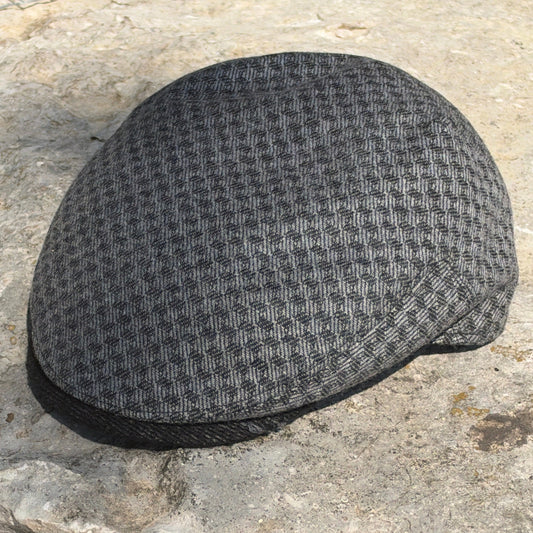 A dark gray and black houndstooth pattern flat cap resting on a textured stone surface.