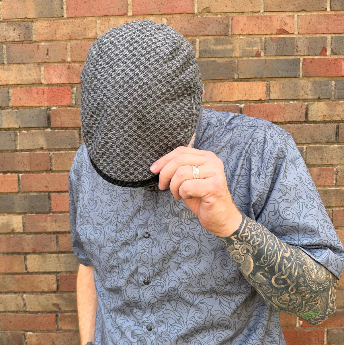 A man wearing a gray and black patterned flat cap and a blue patterned shirt, holding the brim of the cap with his hand in front of a brick wall.
