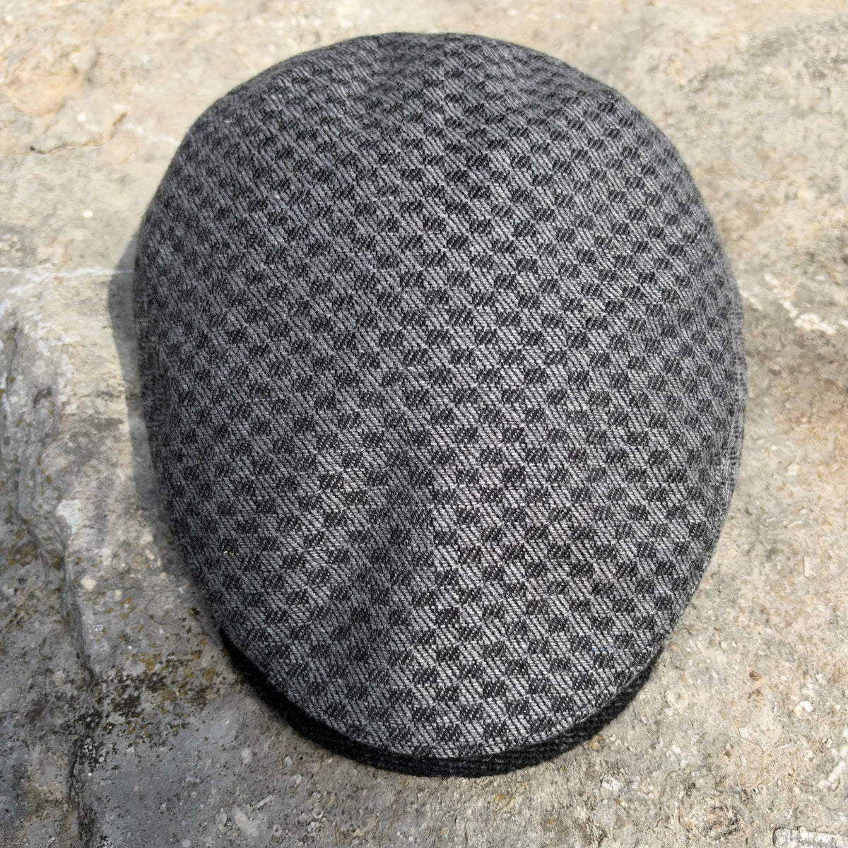 A gray and black textured flat cap resting on a natural stone surface.