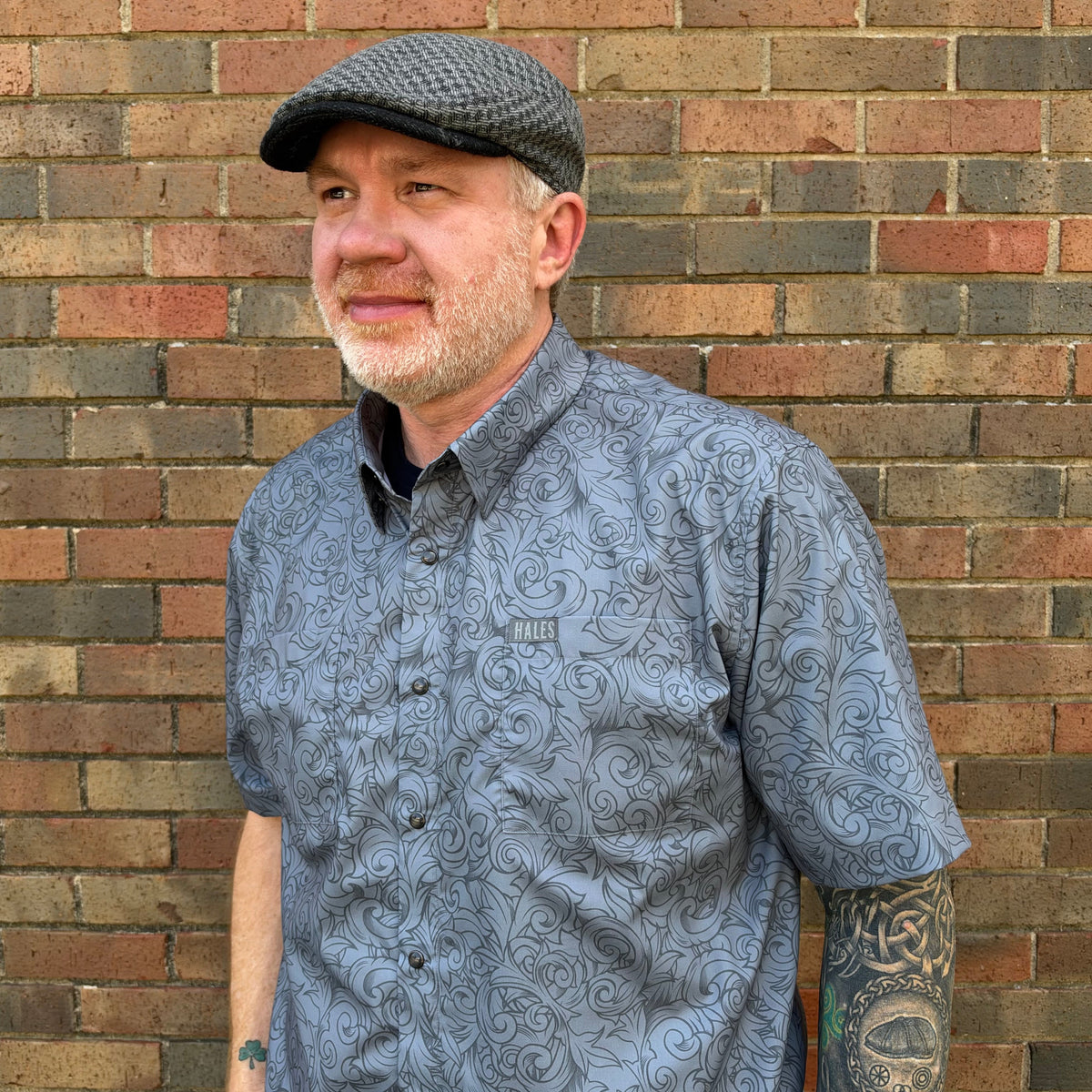 A man with a gray beard and a gray patterned flat cap stands against a brick wall, wearing a gray short-sleeve button-up shirt with a swirling pattern.