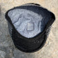 A black and gray patterned flat cap shown from a top-down angle, resting on a textured stone surface. The interior features a gray lining with a black swirl pattern and a central brand patch reading "Graver".