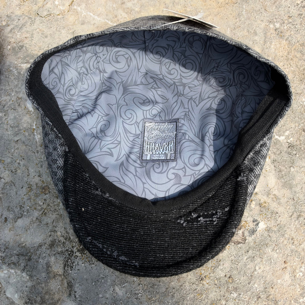 A black and gray patterned flat cap shown from a top-down angle, resting on a textured stone surface. The interior features a gray lining with a black swirl pattern and a central brand patch reading "Graver".