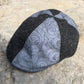 A structured flat cap with a black and gray patterned design, resting on a textured stone surface.