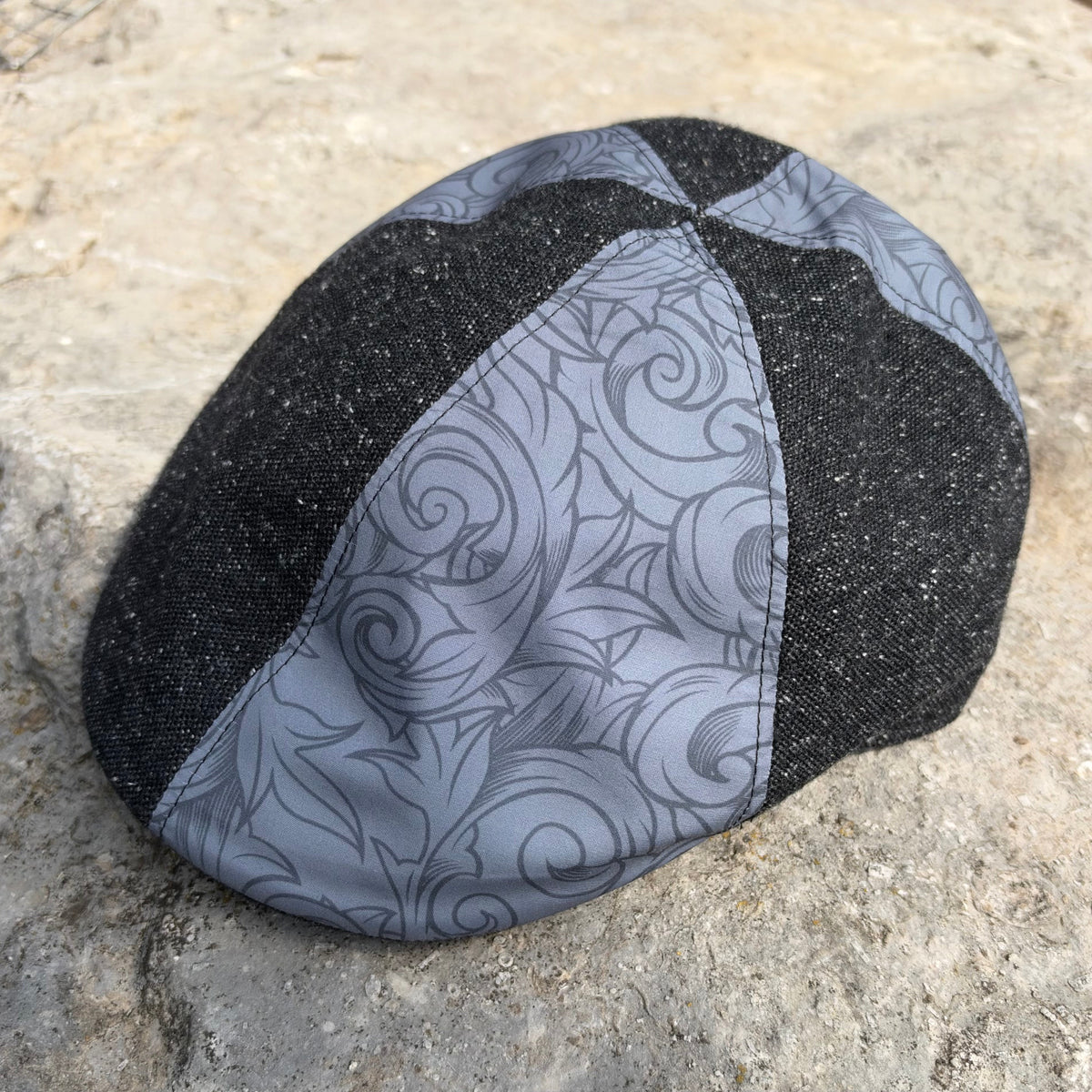 A structured flat cap with a black and gray patterned design, resting on a textured stone surface.