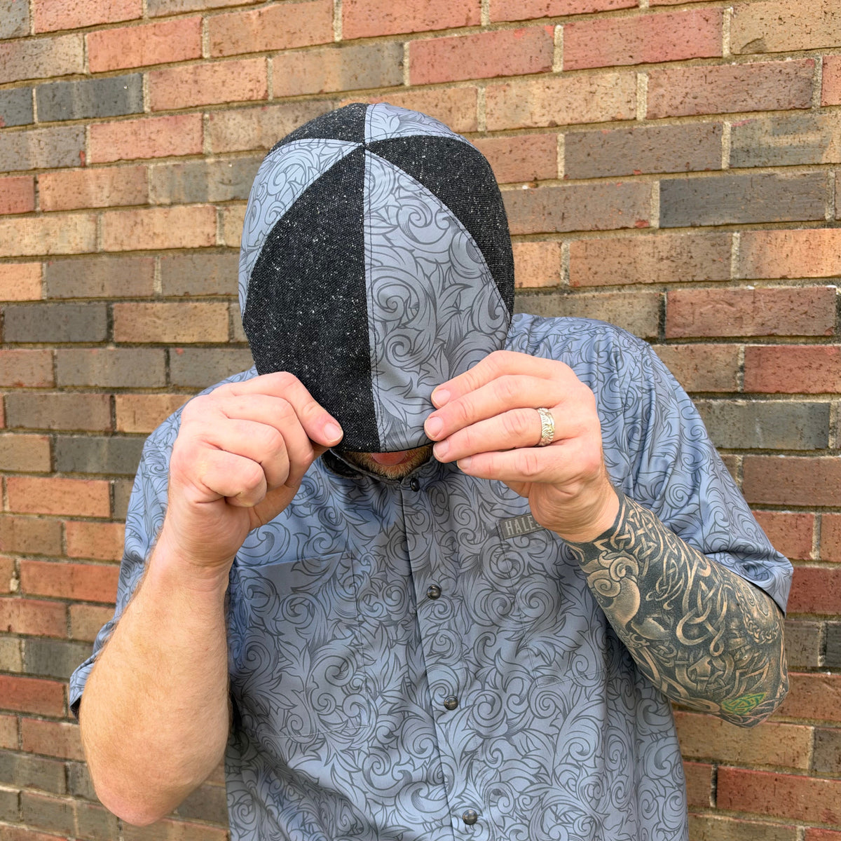 A man wearing a gray and black patterned hood with his hands covering his face, standing in front of a brick wall.