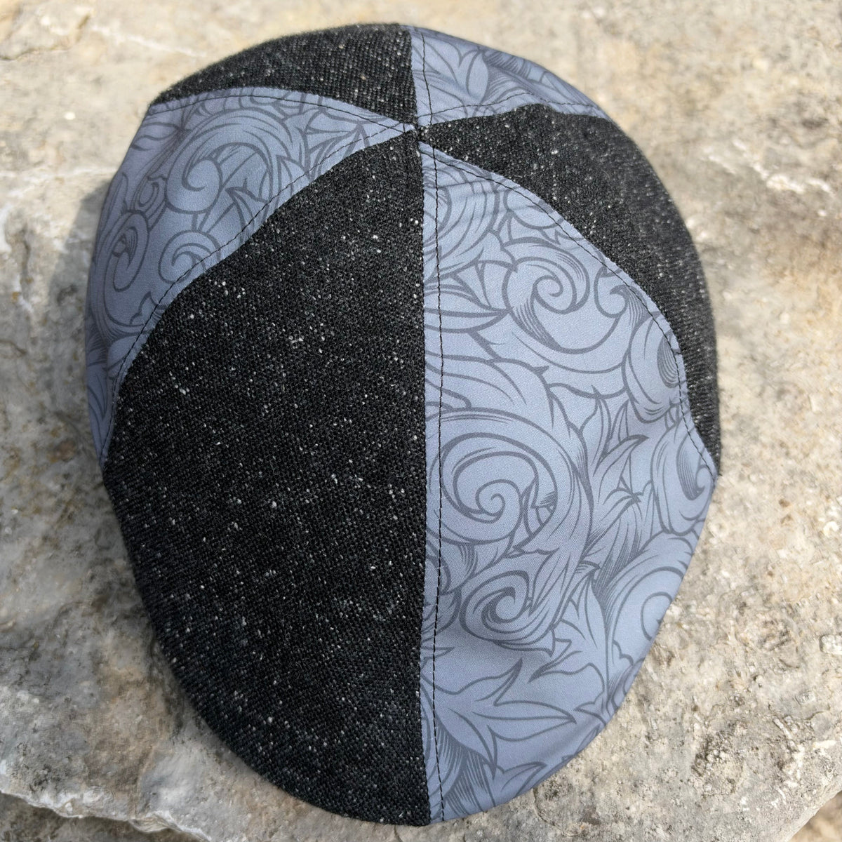 A fabric skullcap with a split design, featuring one side in black with tiny white speckles and the other in light gray with a dark swirl pattern, resting on a textured stone surface.