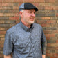 A man wearing a gray short-sleeve button-up shirt with a dark gray swirl pattern and a matching gray flat cap, standing in front of a brick wall.