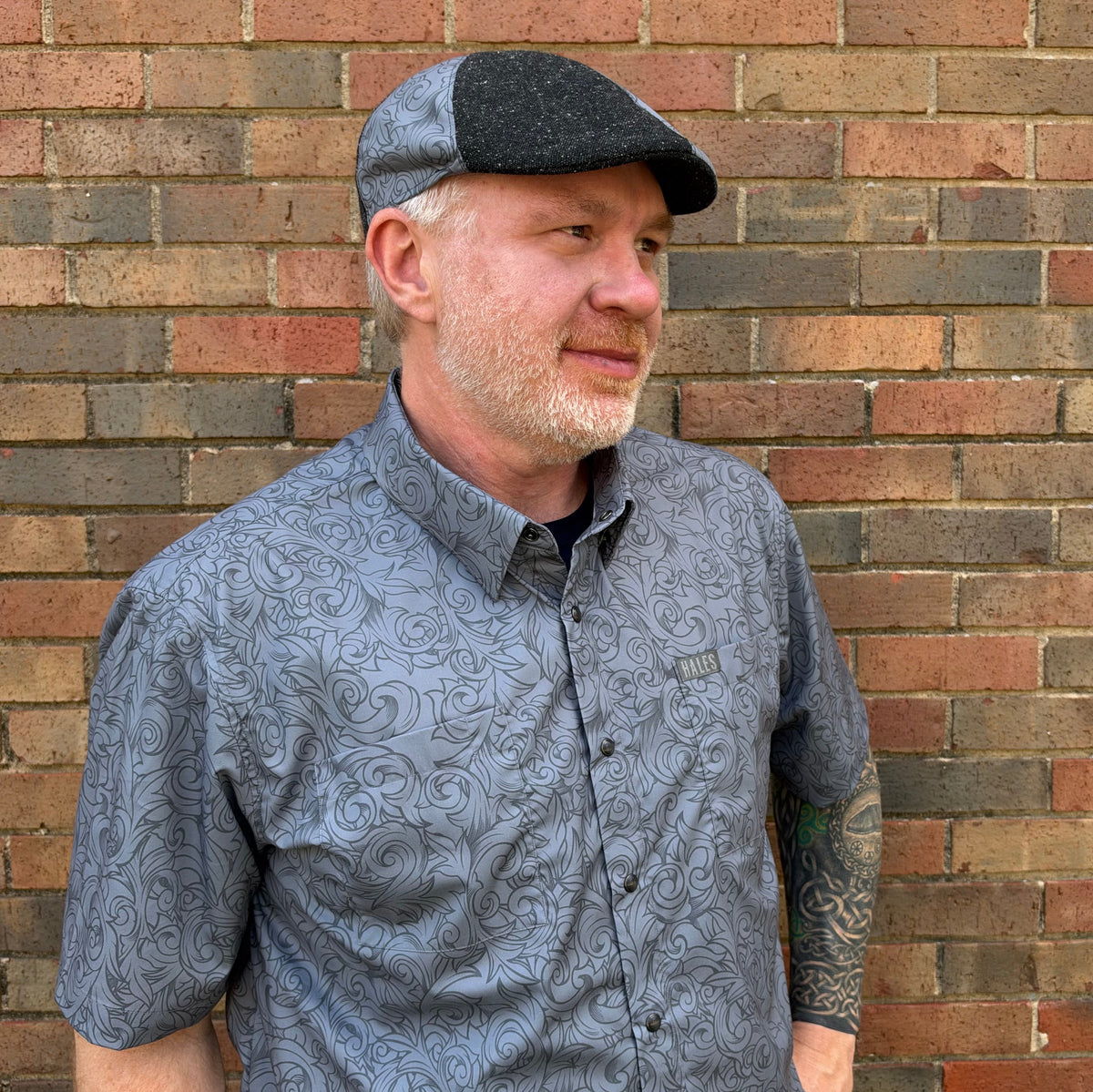 A man wearing a gray short-sleeve button-up shirt with a dark gray swirl pattern and a matching gray flat cap, standing in front of a brick wall.