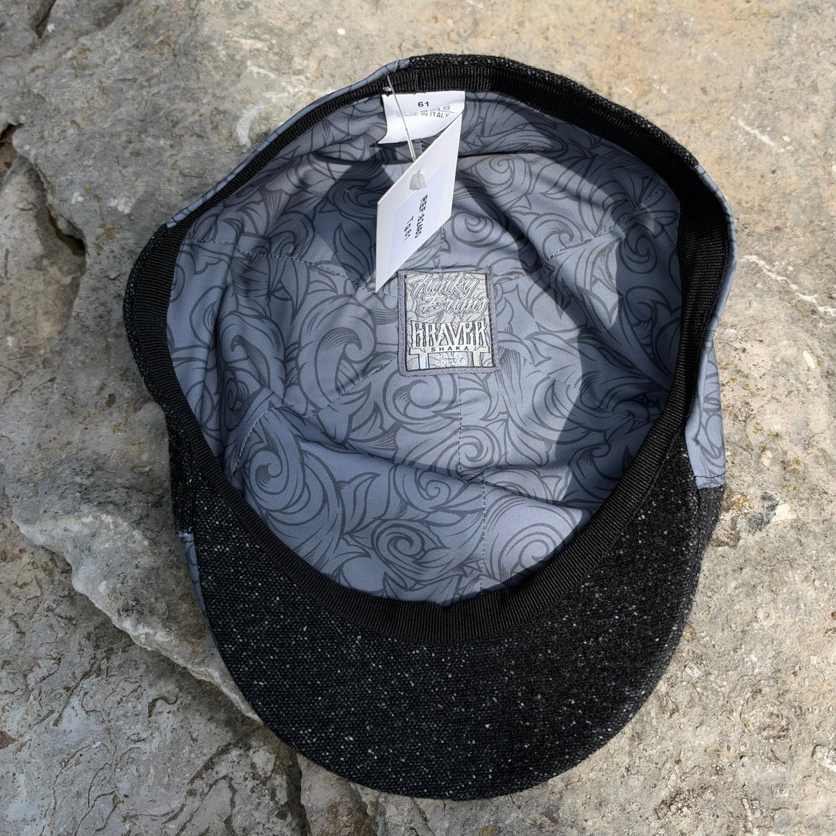 A black baseball cap with a gray patterned interior lining, laid flat on a textured stone surface. The brand name 'GRAVER' is visible on the inner patch.