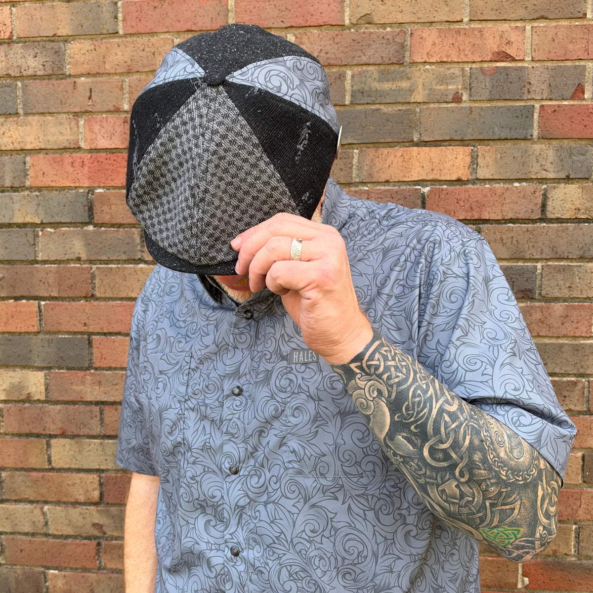 A man wearing a gray and black patterned newsboy cap and a matching gray patterned short-sleeve button-up shirt, standing in front of a brick wall.