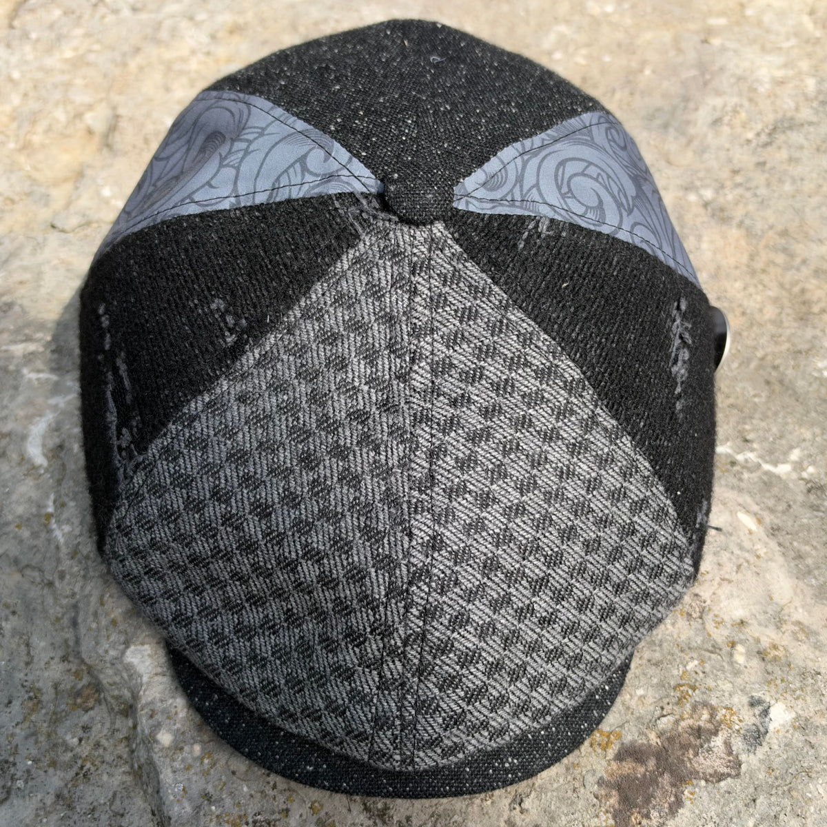 A structured flat cap hat with a black and gray houndstooth pattern on the front and brim, and light gray patterned side panels, resting on a textured stone surface.
