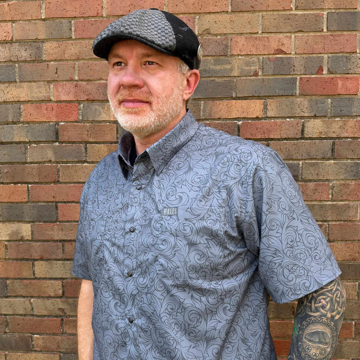A man wearing a gray short-sleeve button-up shirt with a black swirl pattern and a black-and-gray flat cap, standing against a brick wall.