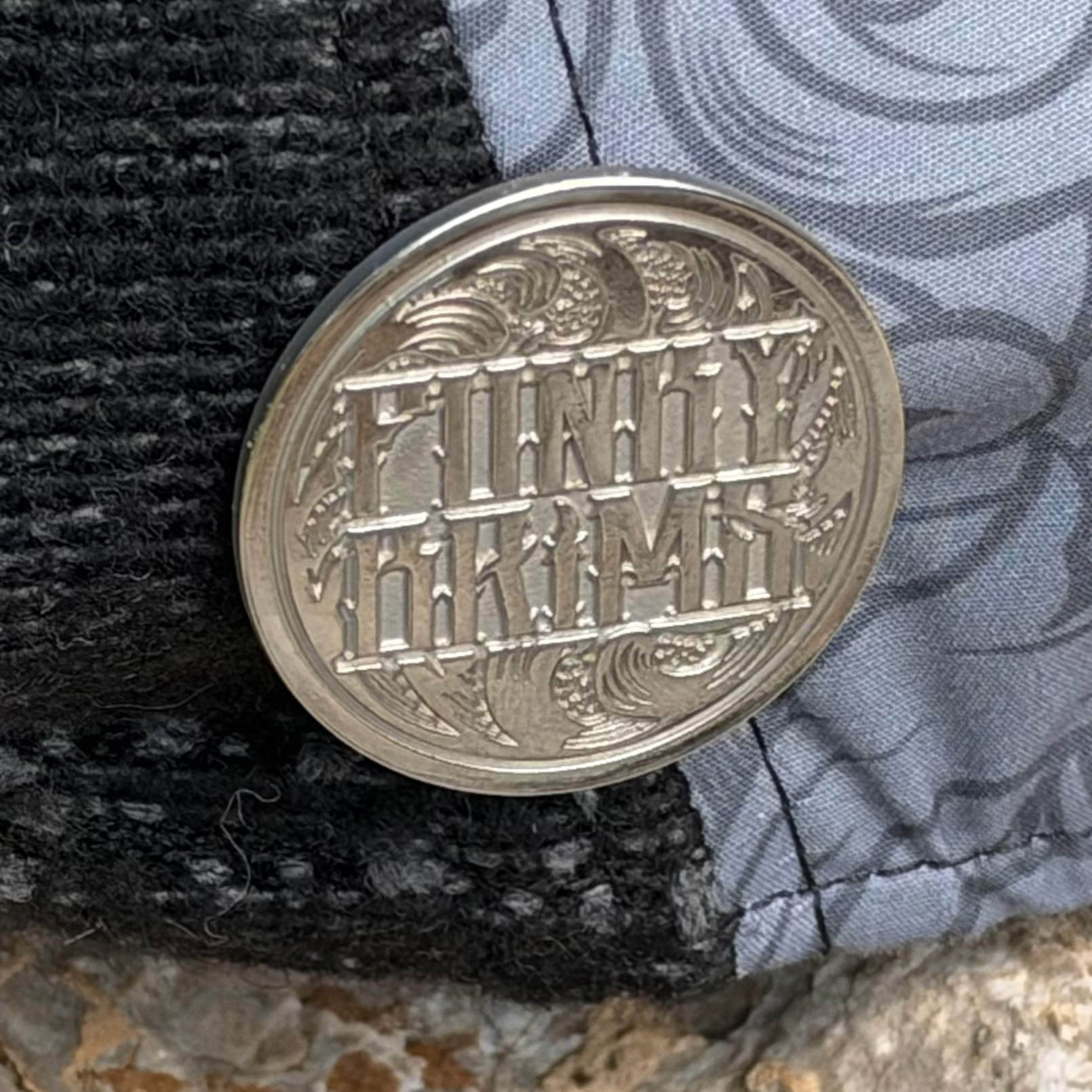 A round silver-toned metal coin or medallion with raised engraved lettering reading 'FINLY TIKIMY' and decorative wave motifs, resting on a black knit fabric next to a gray patterned textile.