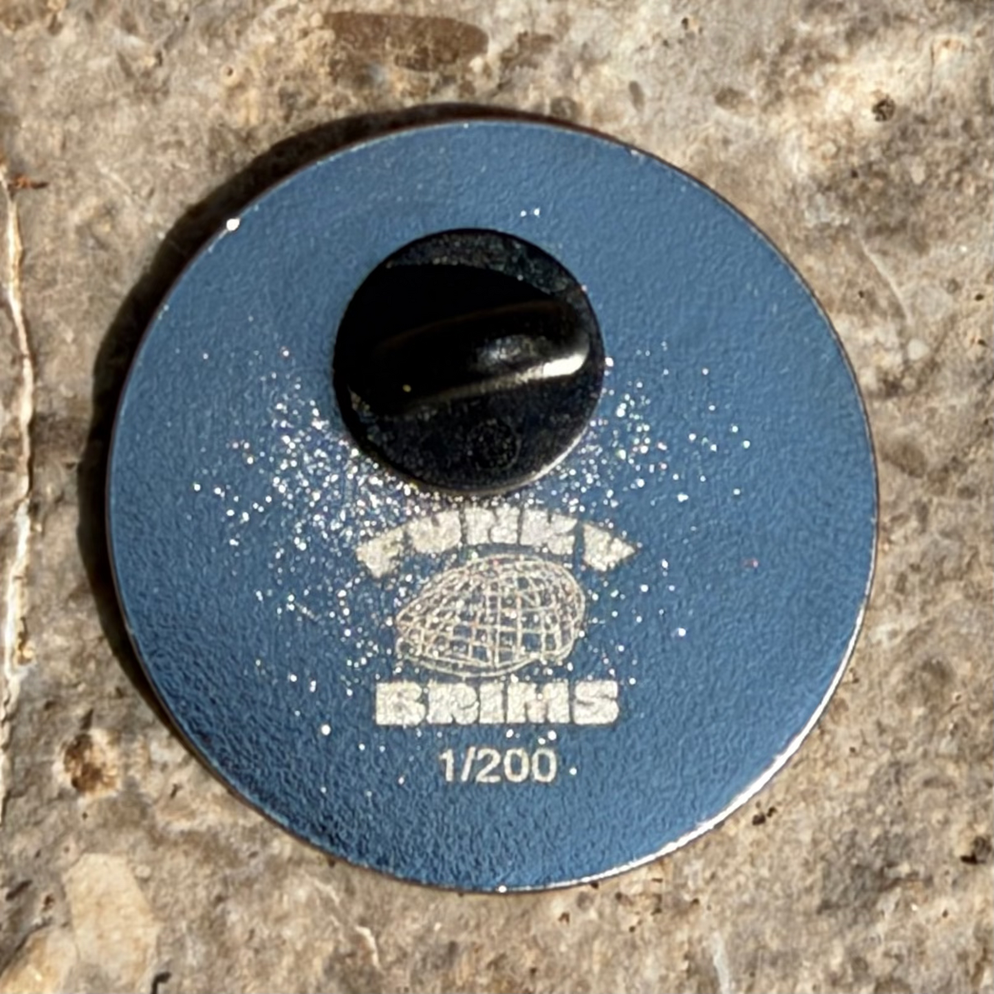 A round blue model kit pinback badge with a black center hole, featuring the text 'FUNNYBRIMS 1/200' and a globe graphic, photographed on a textured stone surface.