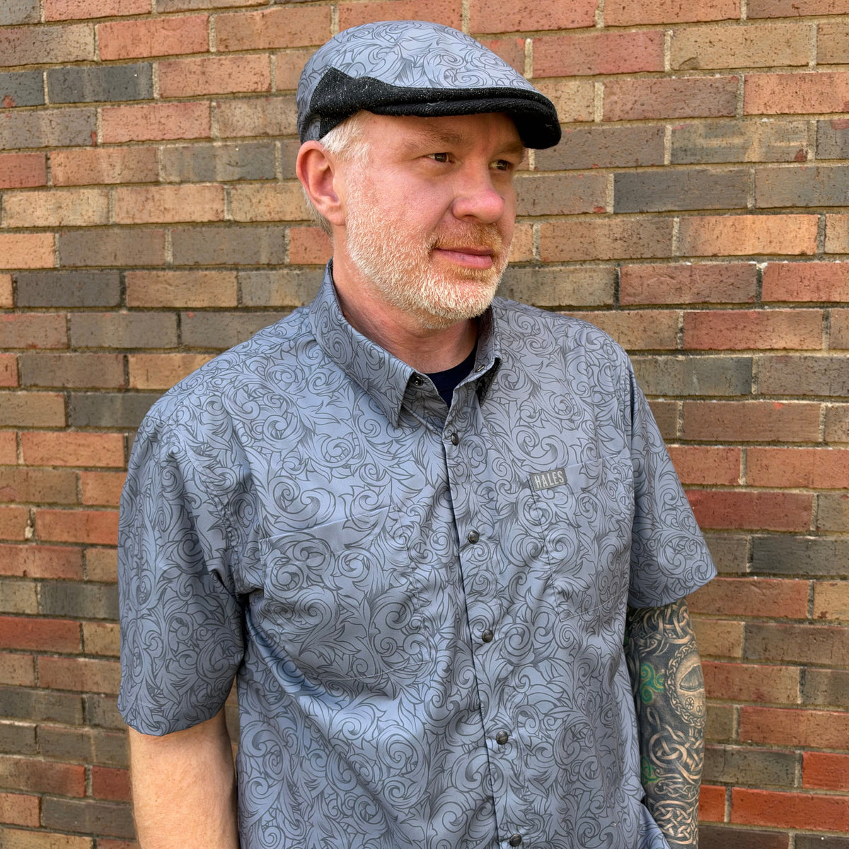 A man wearing a gray short-sleeve button-up shirt with a dark gray scroll pattern and a matching flat cap, standing in front of a red and brown brick wall.
