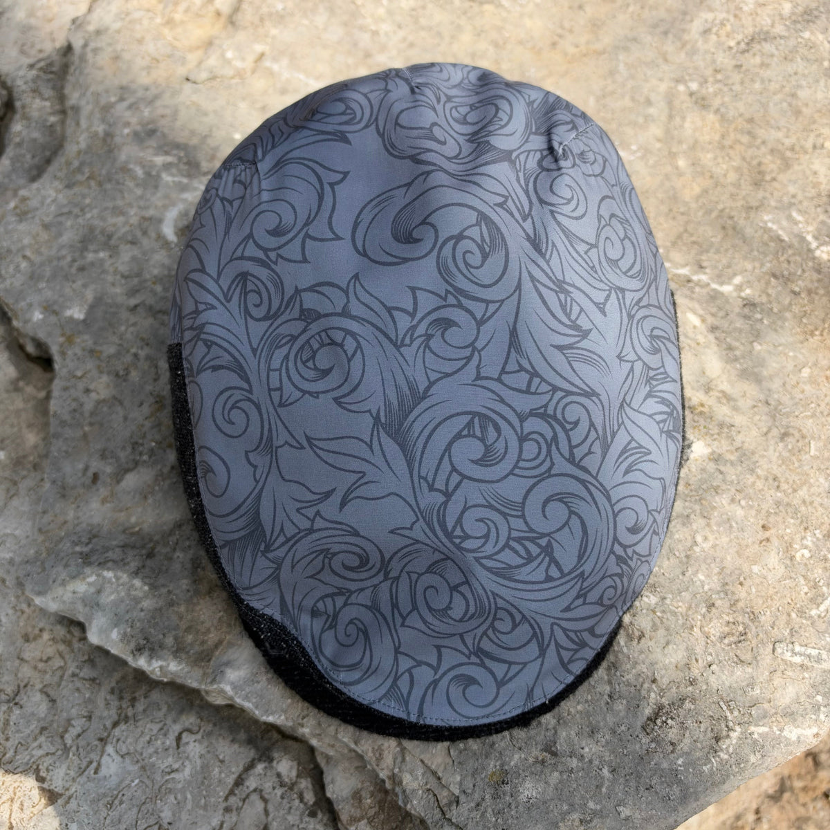 A gray protective helmet cover with a black floral scroll pattern, resting on a textured gray stone surface.