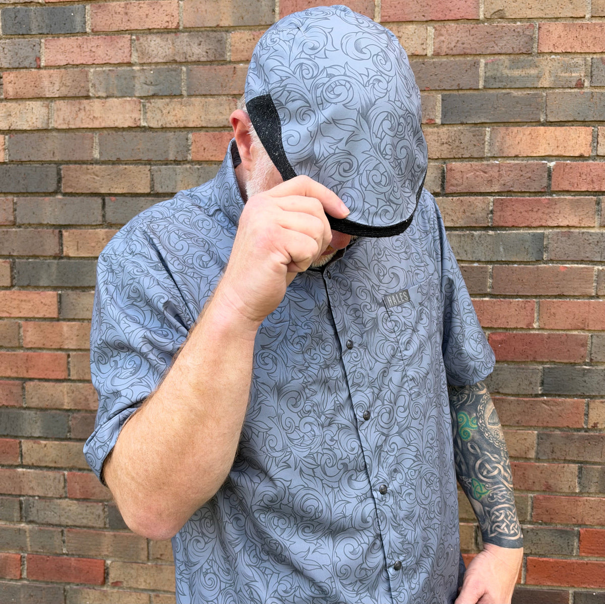 A man wearing a light blue patterned button-down shirt and a matching light blue hooded cap with a black trim, posing in front of a brick wall.