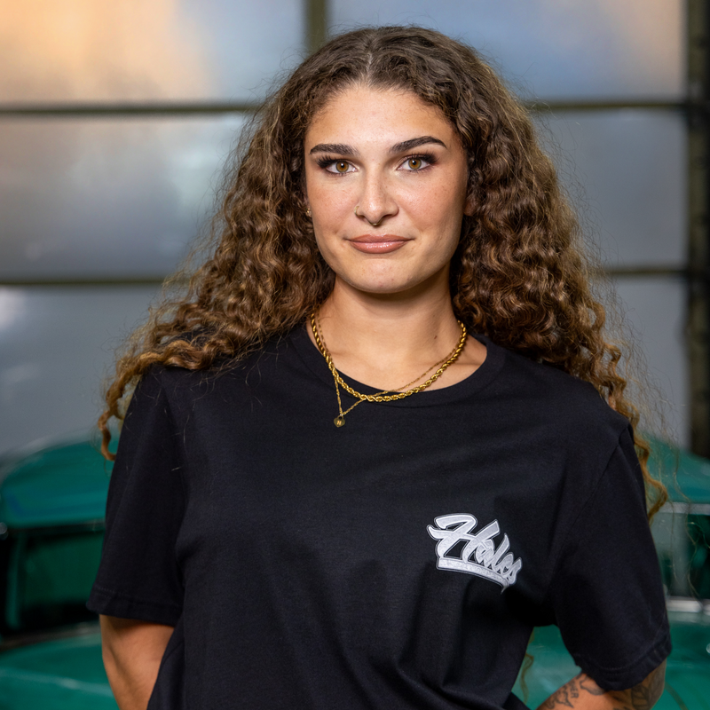 A woman with long curly brown hair wearing a black t-shirt with a white 'Halo' logo on the left chest, standing in front of a teal classic car.