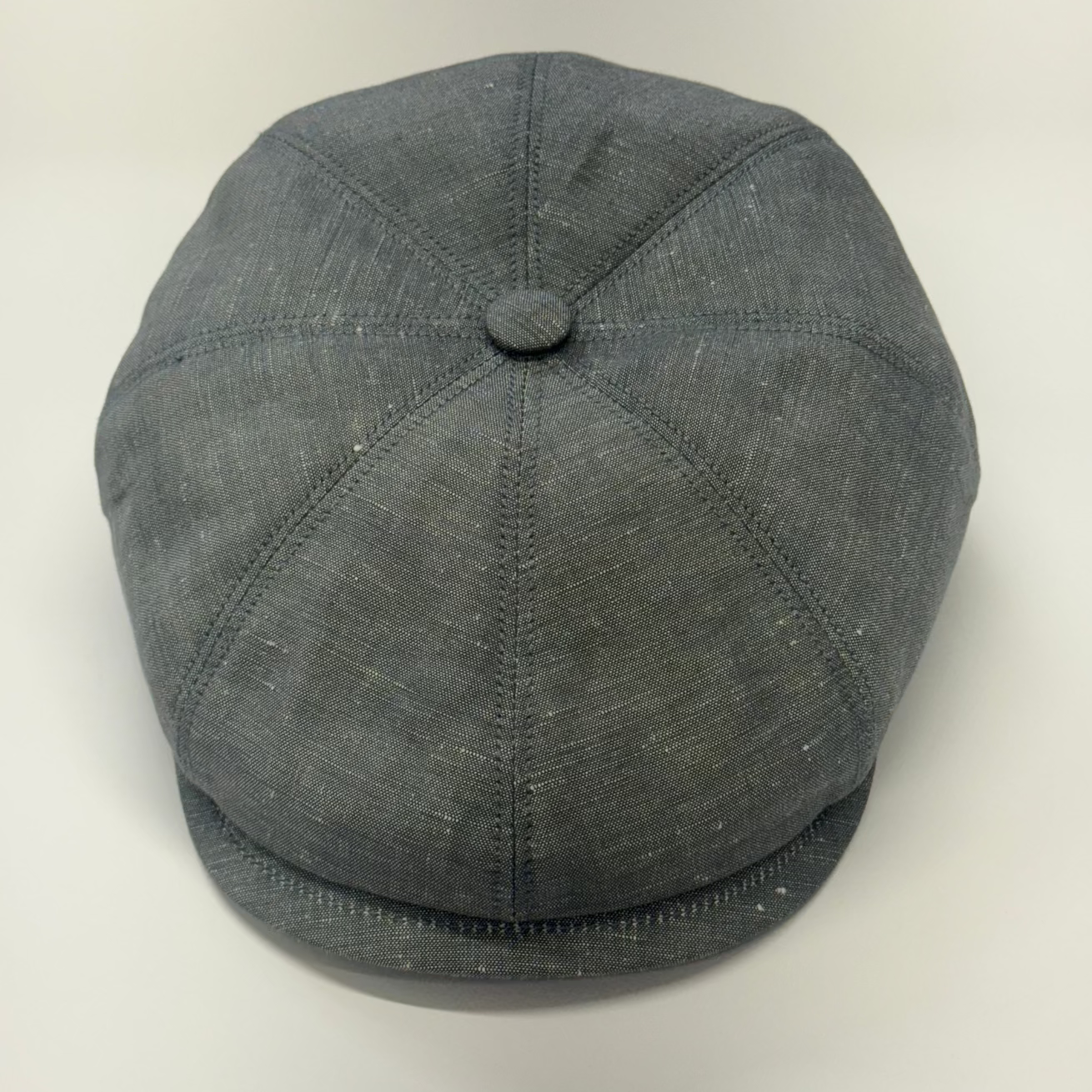 A dark gray textured flat cap with a button on top, photographed from a top-down angle on a light off-white background.