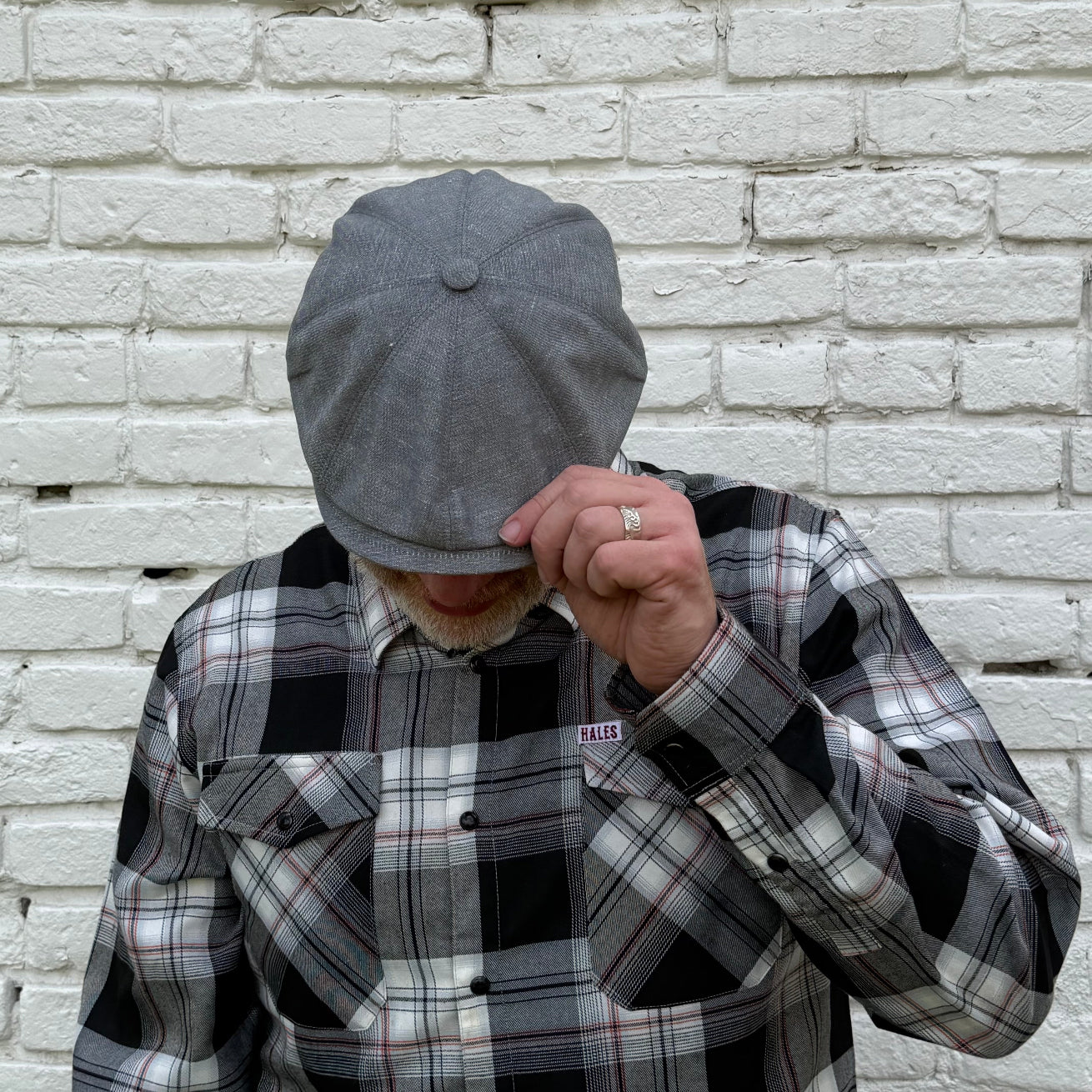 A man wearing a gray newsboy cap and a black, white, and gray plaid flannel shirt stands against a white brick wall, covering his face with the cap while holding his hand up near his face.