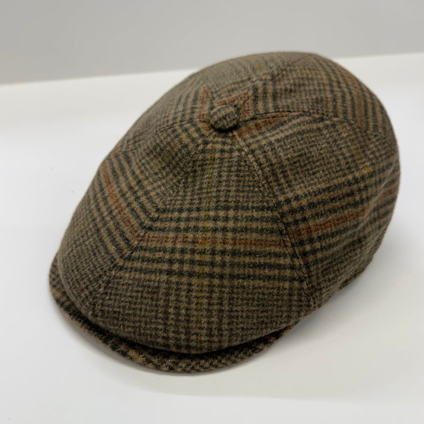 A classic flat cap in a brown and beige houndstooth pattern, photographed on a clean white surface with a neutral gray background.