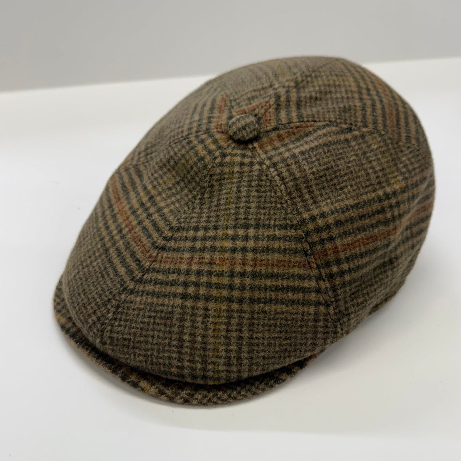 A classic flat cap in a brown and beige houndstooth pattern, photographed on a clean white surface with a neutral gray background.