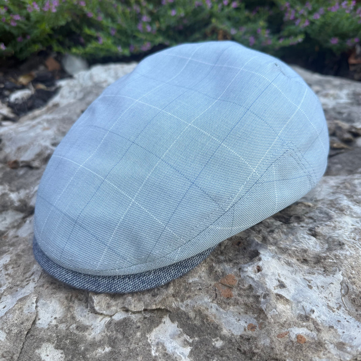 A light blue flat cap with a subtle windowpane check pattern and a darker gray ribbed brim, resting on a textured stone surface with blurred green foliage in the background.