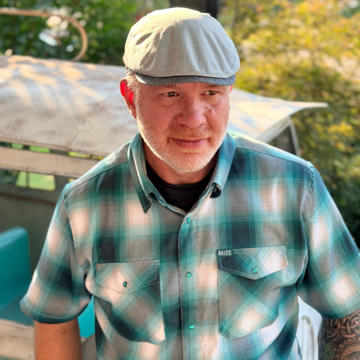 A man wearing a teal, green, and gray plaid short-sleeve button-up shirt and a light gray flat cap, standing outdoors in front of a rustic vehicle.