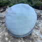 A light blue-gray flat cap with a subtle white windowpane check pattern, resting on a textured stone surface with blurred green foliage in the background.