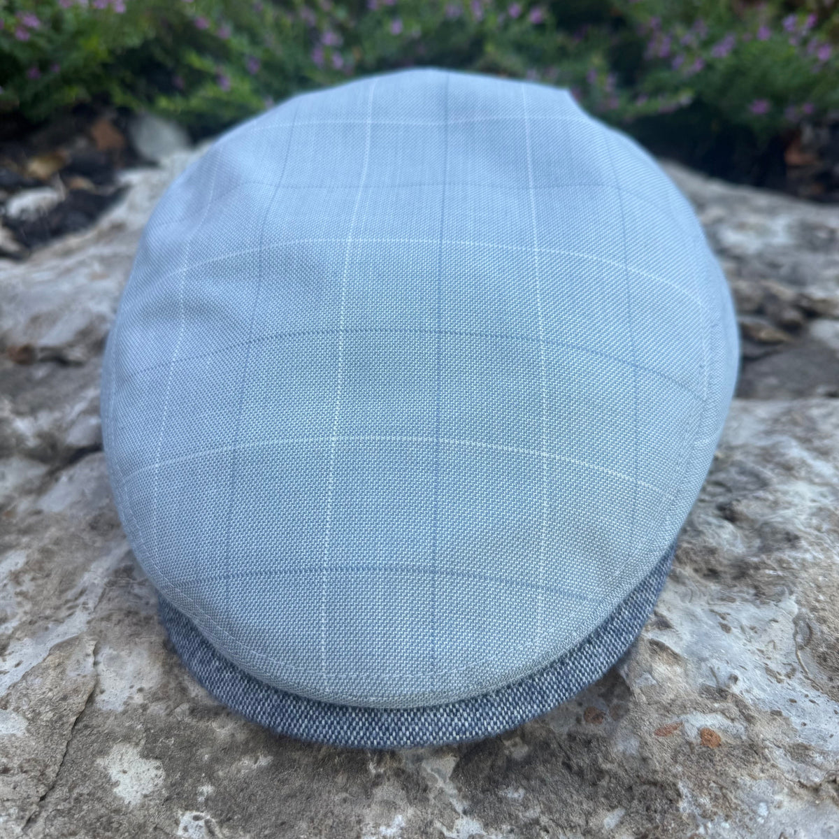 A light blue-gray flat cap with a subtle white windowpane check pattern, resting on a textured stone surface with blurred green foliage in the background.
