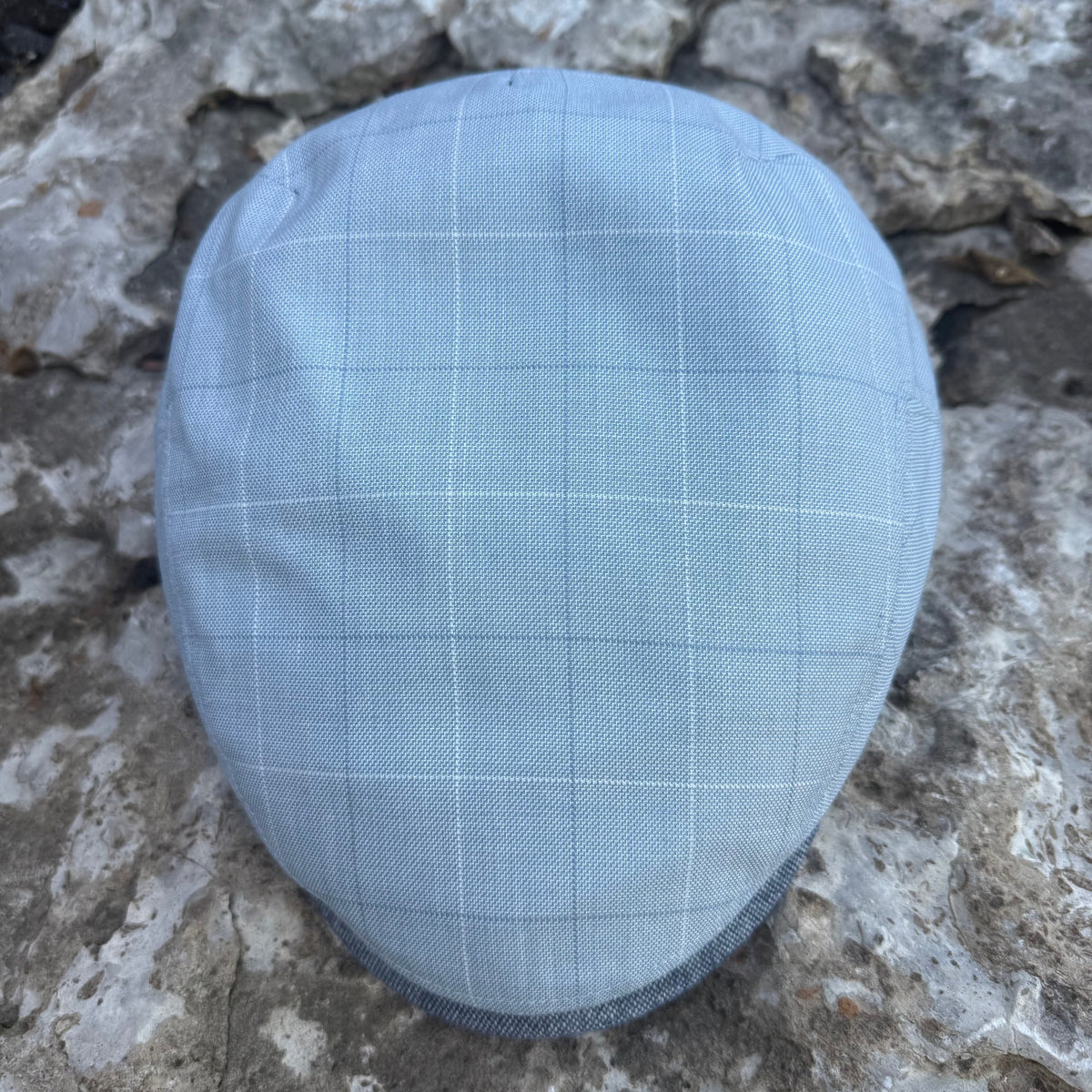 A light blue flat cap with a subtle white windowpane check pattern, photographed from above on a textured gray stone surface.
