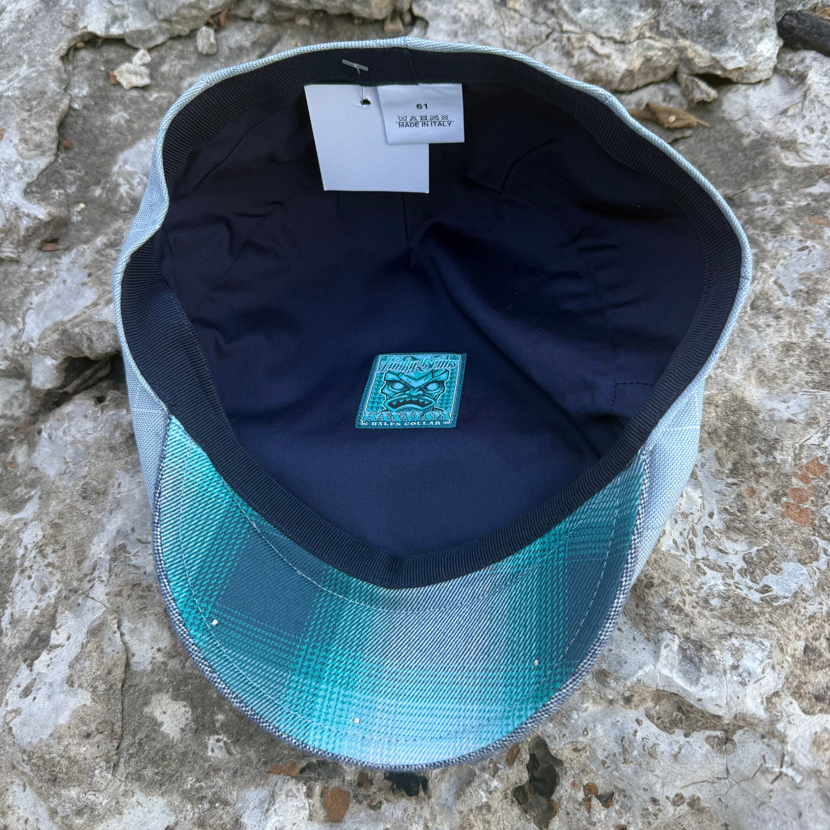A baseball cap photographed from above on a rough stone surface. The cap has a dark navy blue interior, a white hangtag, and a front brim with a teal-to-light-blue gradient mesh pattern.