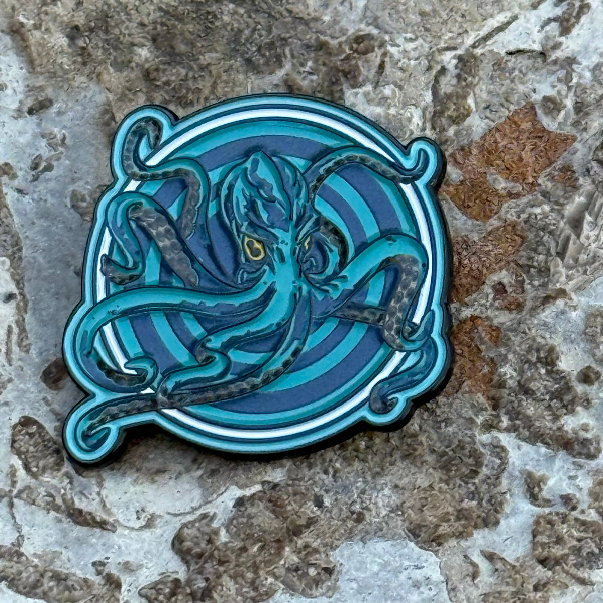 An enamel pin with a  blue and teal Octopus over concentric circles.