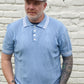 Man wearing a light blue polo shirt and jeans standing against a white brick wall.