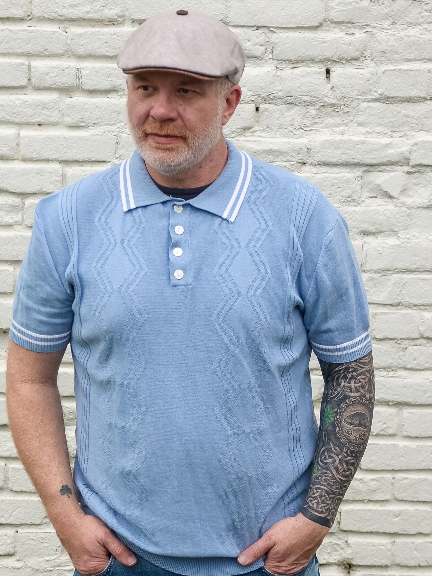 Man wearing a light blue polo shirt and jeans standing against a white brick wall.