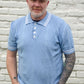 Man wearing a blue polo shirt and jeans standing against a light brick wall.