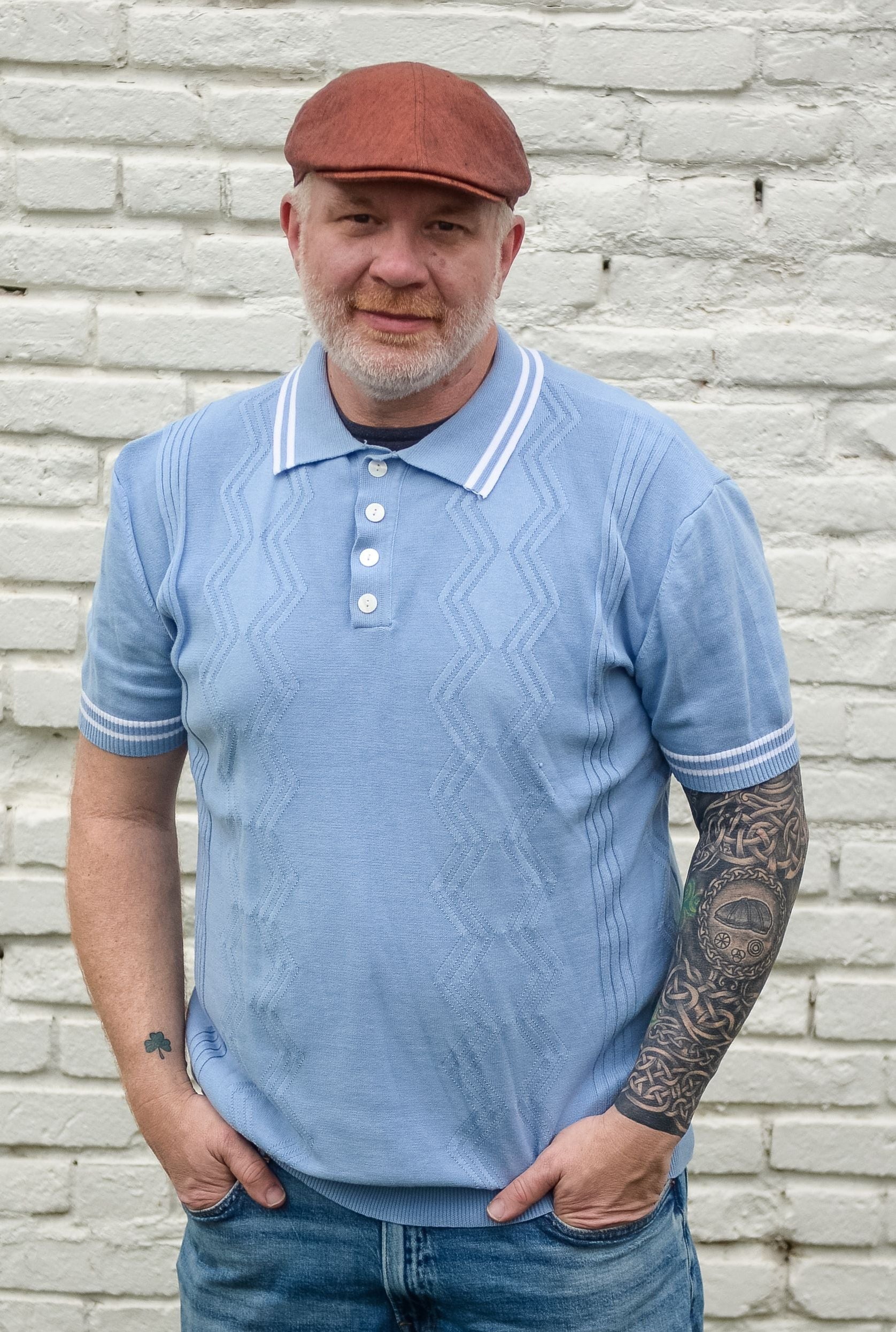 Man wearing a blue polo shirt and jeans standing against a light brick wall.