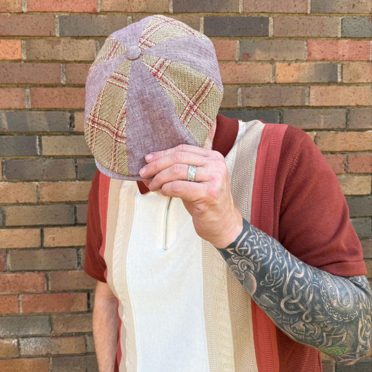 A person wearing a textured tweed flat cap with a muted purple and beige plaid pattern, standing in front of a brick wall.